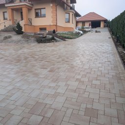 Beżowa kostka brukowa na podjeździe do domu jednorodzinnego z garażem. W tle widoczny fragment ogrodu i budynku. Ułożona kostka tworzy wzór z prostokątów.