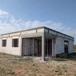 Niedokończony, parterowy dom z białego bloczka, z widocznymi podporami i deskami szalunkowymi, na tle błękitnego nieba z chmurami. Stan surowy otwarty.