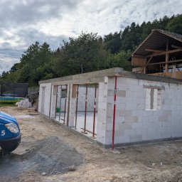 Budowa domu z białych bloczków, widoczne podpory i częściowo wzniesione ściany. W tle las i drewniana konstrukcja. Na pierwszym planie gruz i samochód dostawczy.