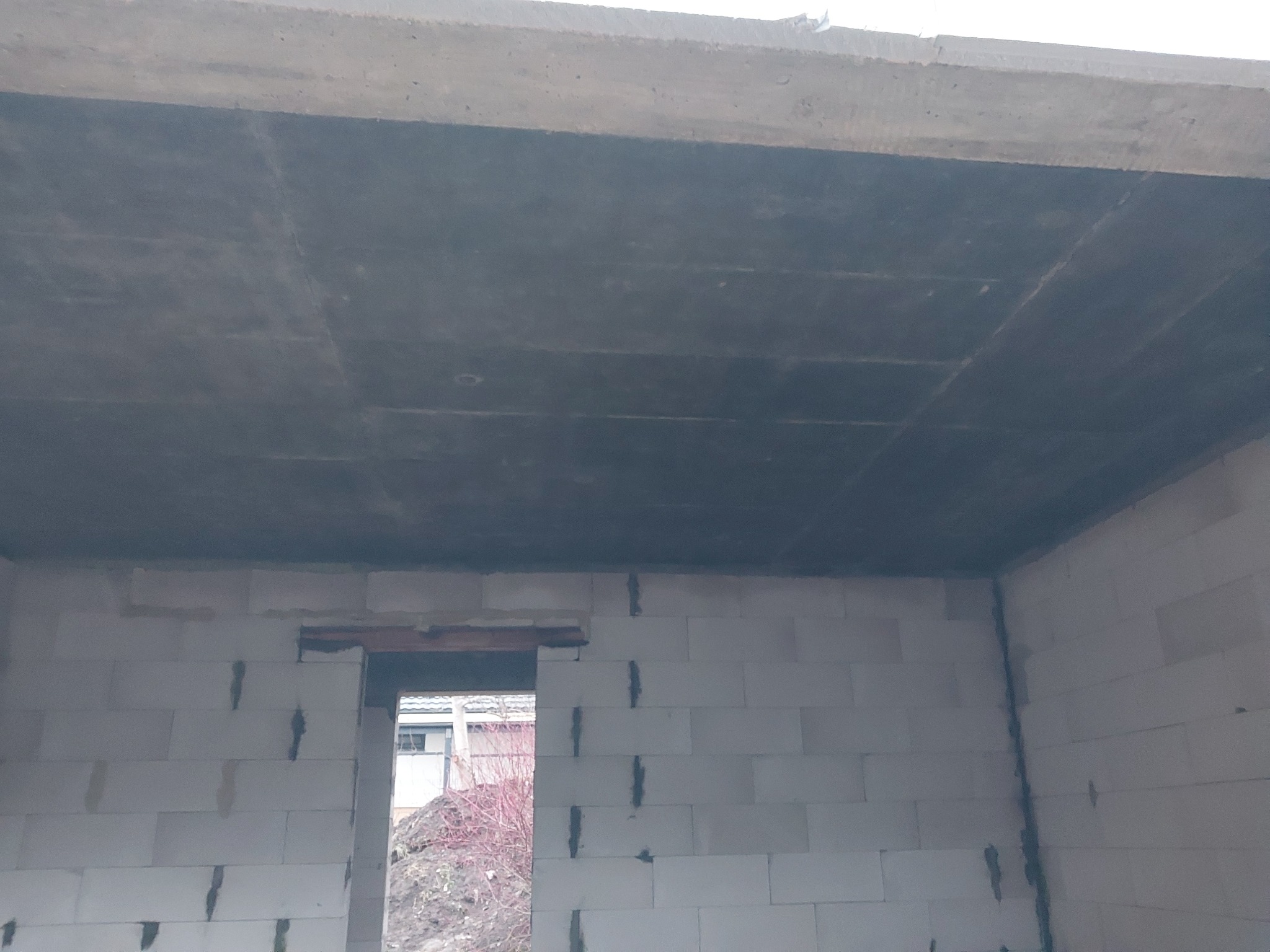 Surowy stan domu w trakcie budowy: widok na betonowy strop i ściany z białych bloczków, z widocznym otworem okiennym i stertą ziemi na zewnątrz.