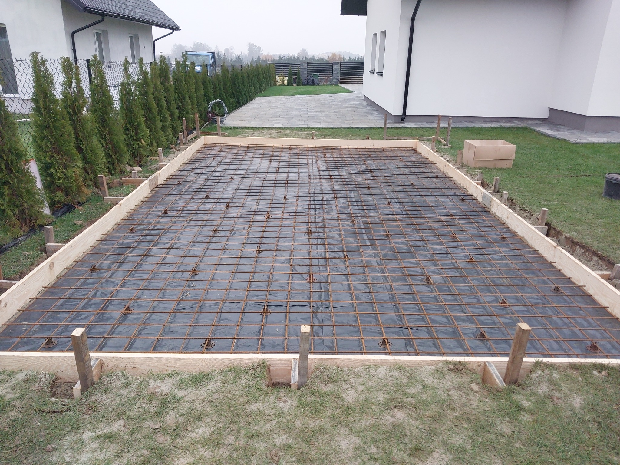 Przygotowanie zbrojenia pod wylewkę fundamentową na gruncie, widoczne drewniane szalunki, folia budowlana i siatka zbrojeniowa, w tle dom jednorodzinny i tuje.