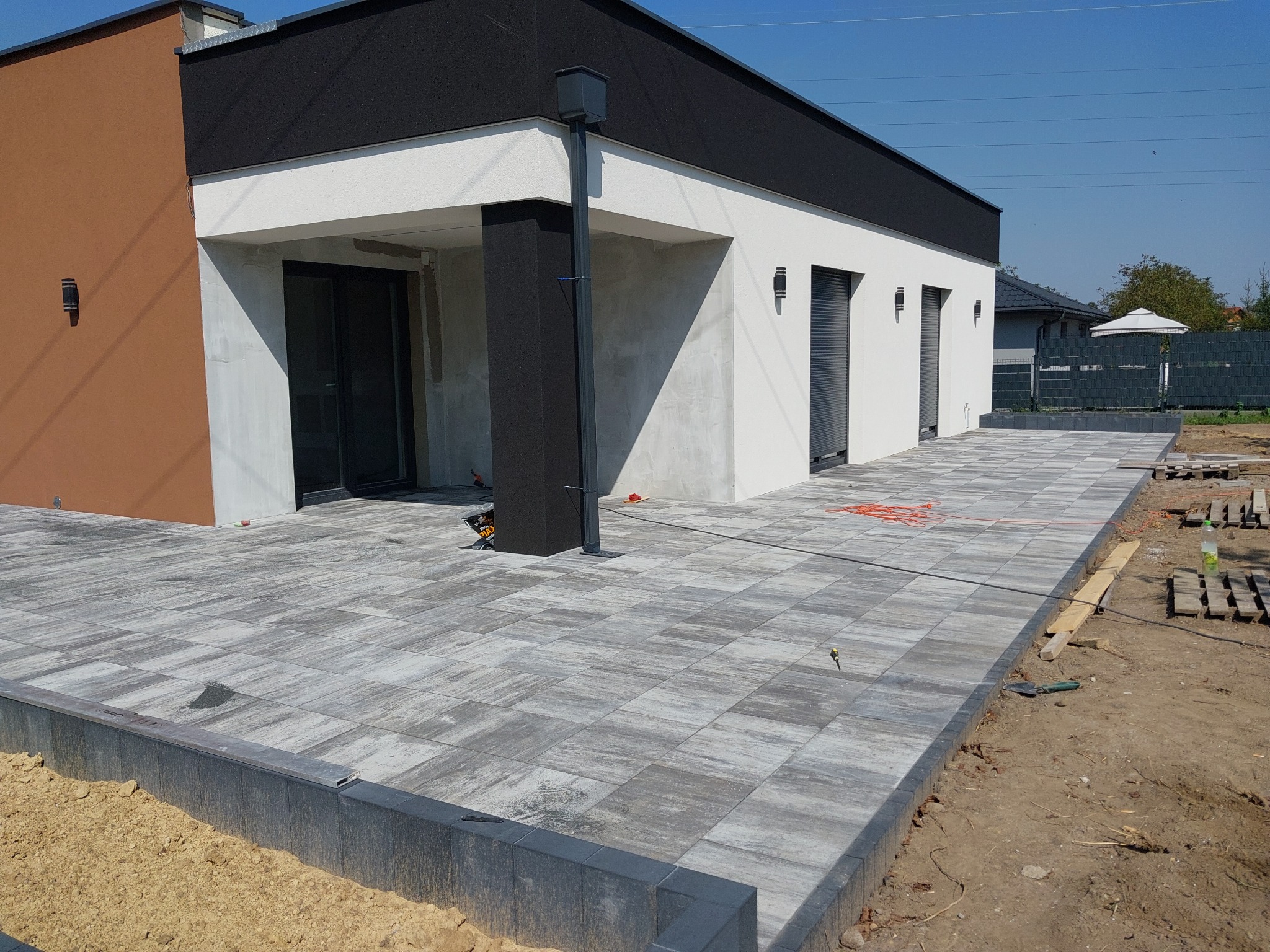 Nowoczesny dom z szarą kostką brukową na tarasie, widoczne złączenia kostki i obrzeża, w tle fragment ogrodzenia panelowego.