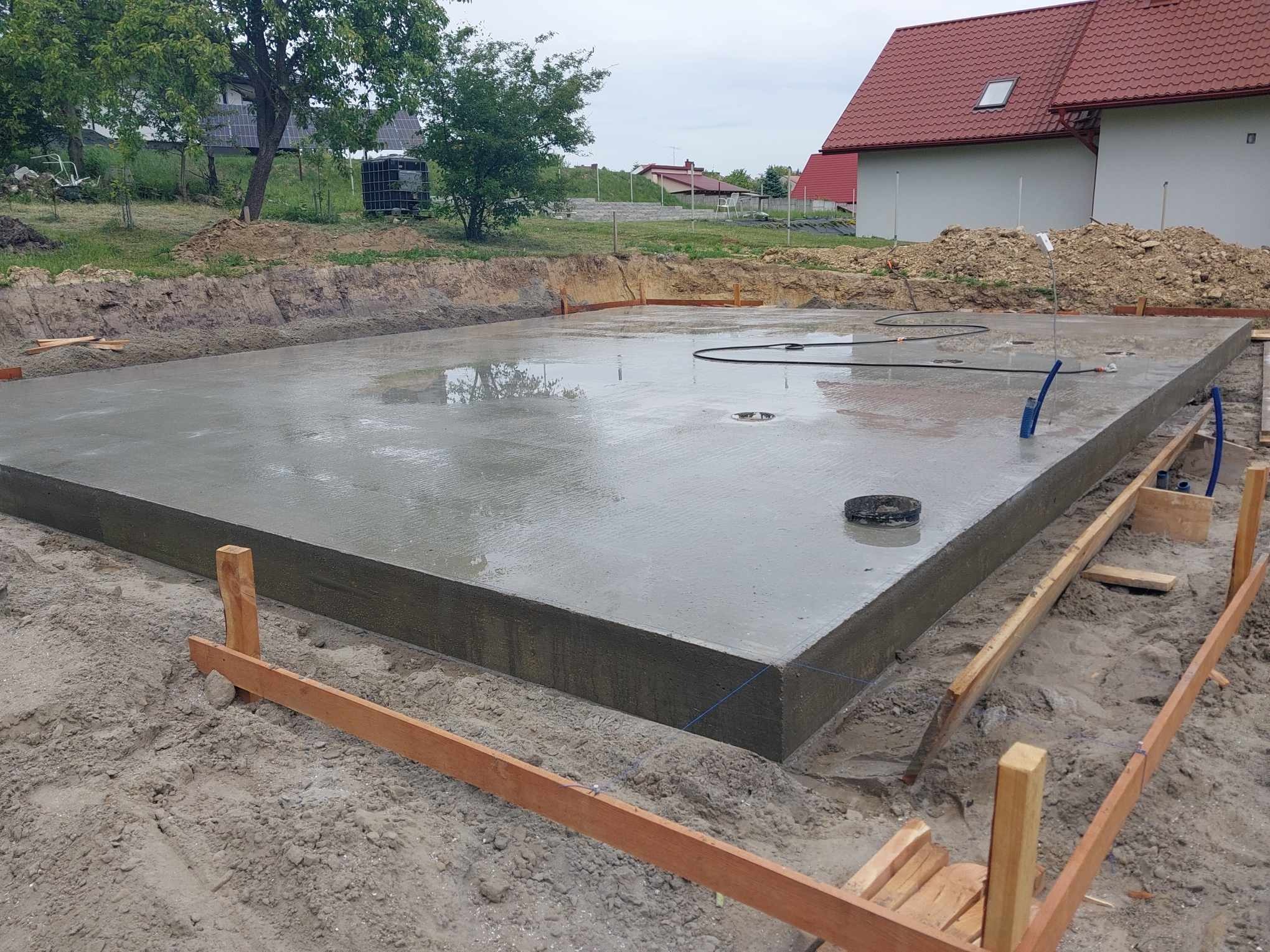 Świeżo wylana, mokra płyta fundamentowa domu jednorodzinnego, otoczona deskowaniem i ziemią, widoczne elementy instalacji w płycie, w tle dom z czerwoną dachówką.
