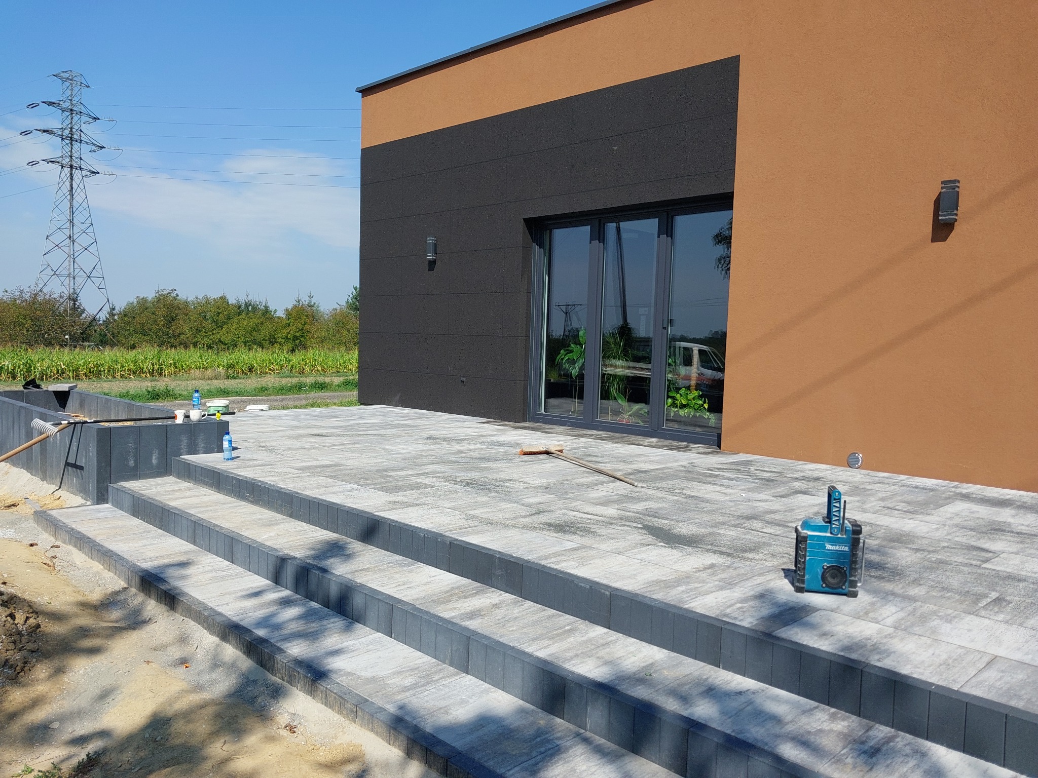 Nowoczesny taras z kostki brukowej w odcieniach szarości, z trzema stopniami prowadzącymi do domu o elewacji w kolorach brązu i grafitu, z widoczną wieżą wysokiego napięcia w tle i radiem...