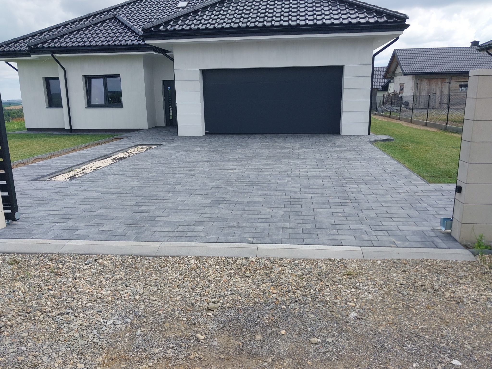 Ułożona szara kostka brukowa przed nowoczesnym domem z garażem; widoczne odwodnienie liniowe z białym grysem, obrzeża betonowe, oraz fragment bramy wjazdowej.