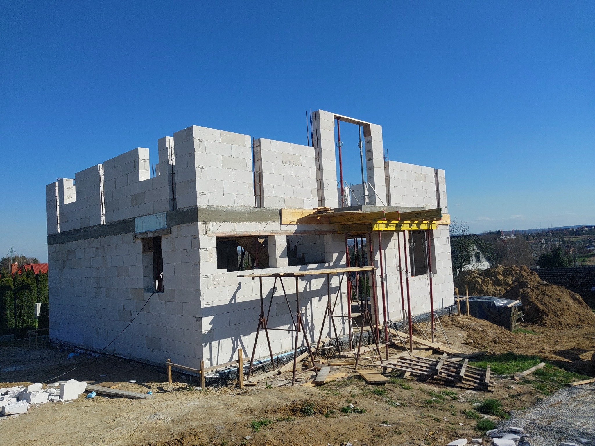 Surowy stan domu jednorodzinnego z białych bloczków, widoczne otwory okienne i drzwiowe, częściowo zadaszony, na pierwszym planie podpory szalunkowe i stos desek, w tle krajobraz podmiejski...