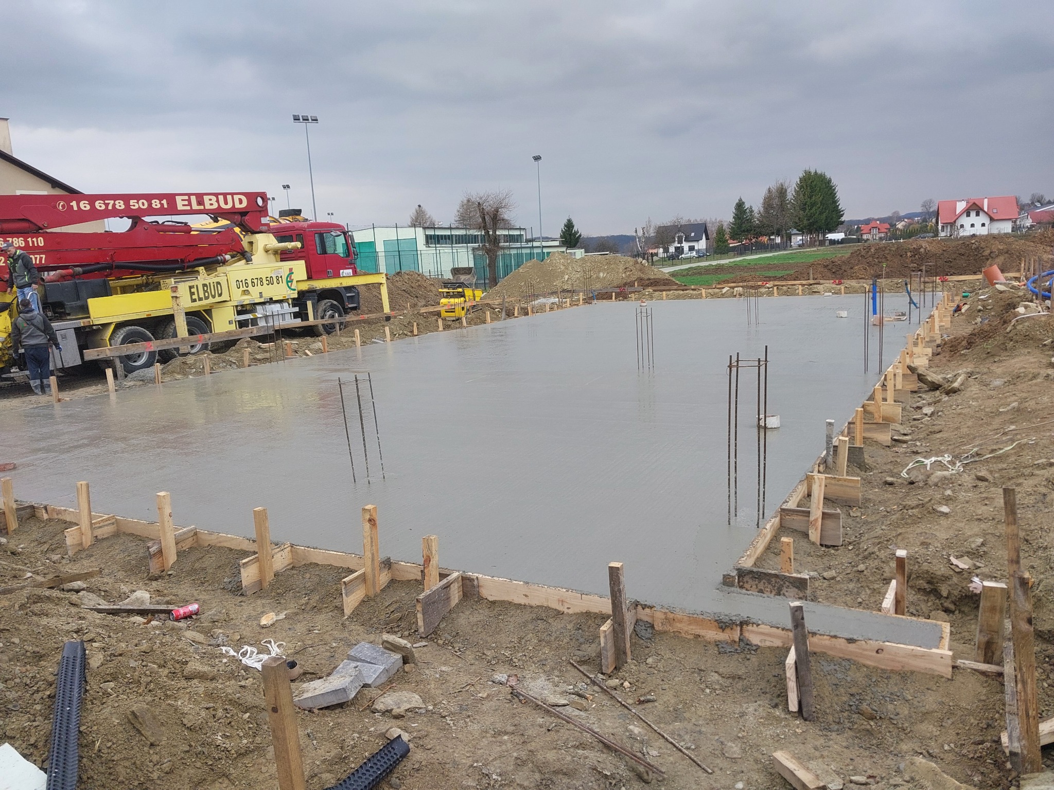 Świeżo wylana, gładka posadzka betonowa w drewnianym szalunku, z wystającymi prętami zbrojeniowymi, z ciężarówką z pompą do betonu Elbud w tle, na placu budowy w pochmurny dzień.