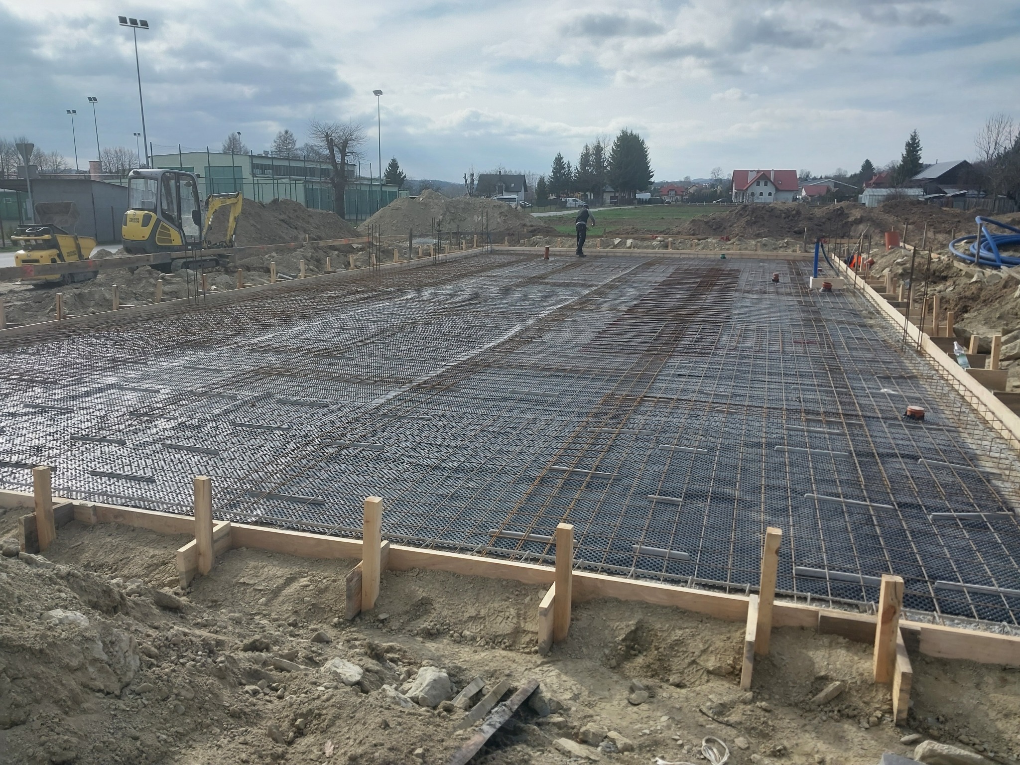 Przygotowanie podłoża pod wylewkę betonową: szalunek z desek, siatka zbrojeniowa i warstwa izolacyjna z folii kubełkowej, widoczny pracownik i sprzęt budowlany w tle.