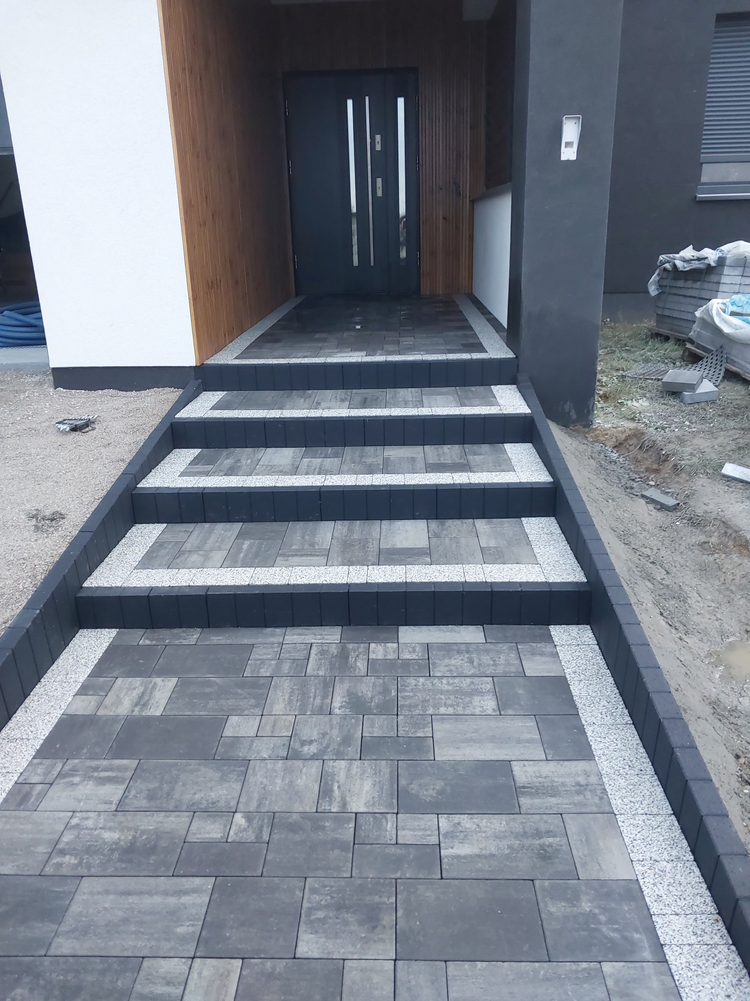 Nowoczesne schody wejściowe wyłożone szarą kostką brukową z dekoracyjnym obramowaniem z drobnego kamienia, prowadzące do drzwi wejściowych budynku.