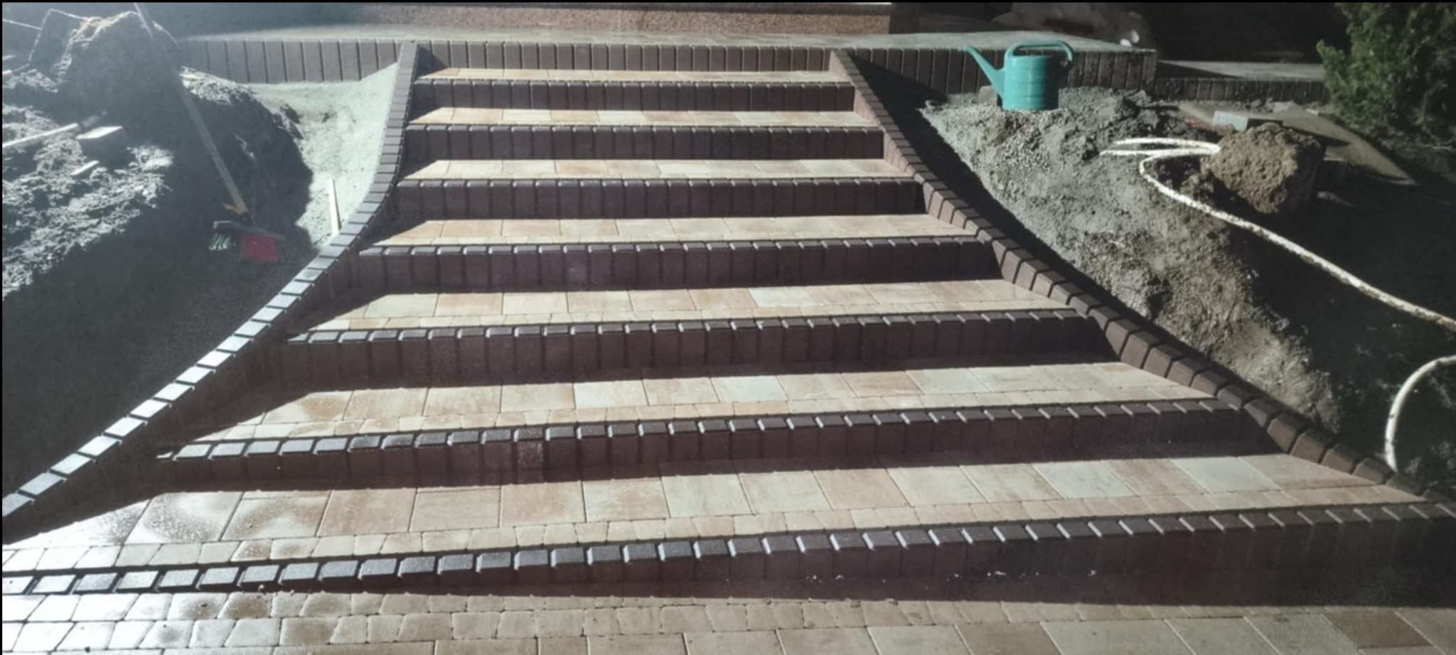 Schody z kostki brukowej w odcieniach beżu i brązu, z widocznym fragmentem terenu przygotowanego pod dalsze prace. W tle zielony konewka i fragment roślinności.