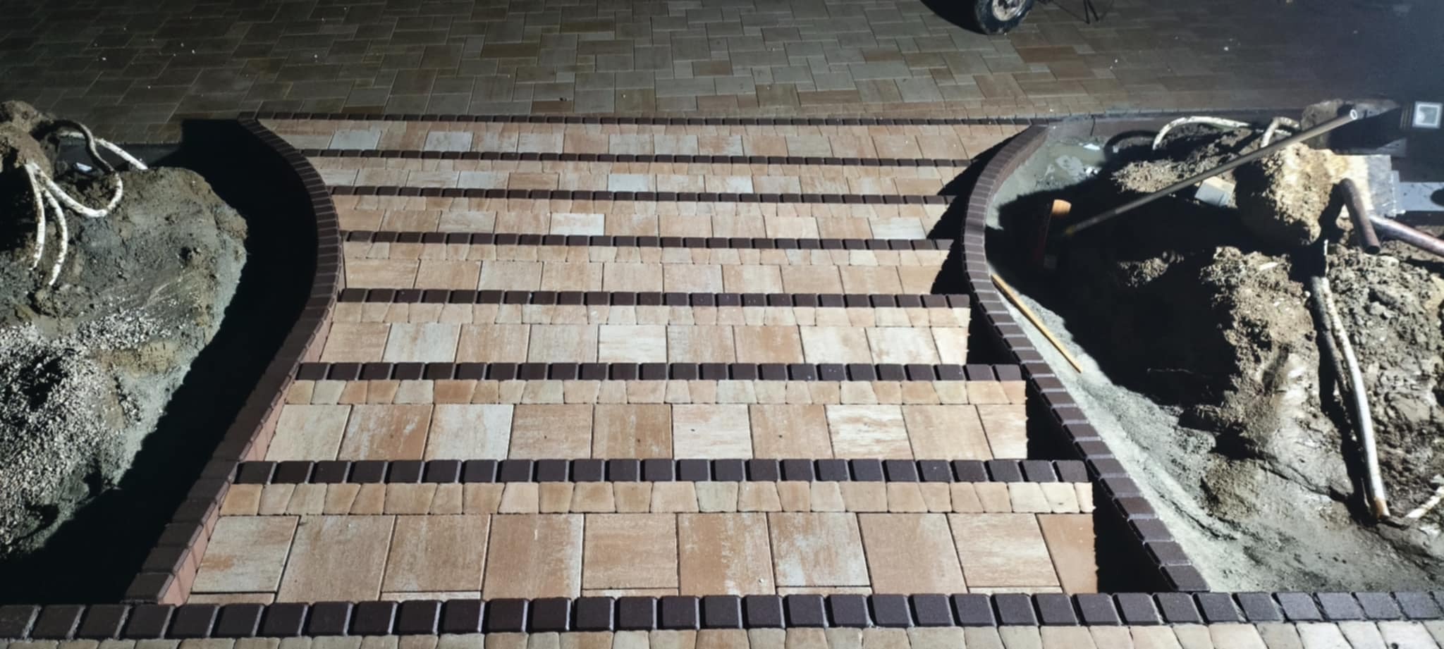 Schody z kostki brukowej w odcieniach beżu i brązu, z elementami dekoracyjnymi. Widoczne przygotowane miejsca na rośliny po bokach. Ujęcie z góry.