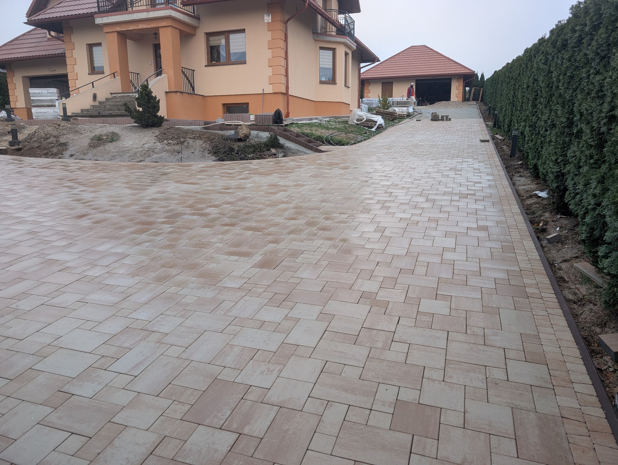 Beżowa kostka brukowa na podjeździe do domu jednorodzinnego z garażem. W tle widoczny fragment ogrodu i budynku. Ułożona kostka tworzy wzór z prostokątów.