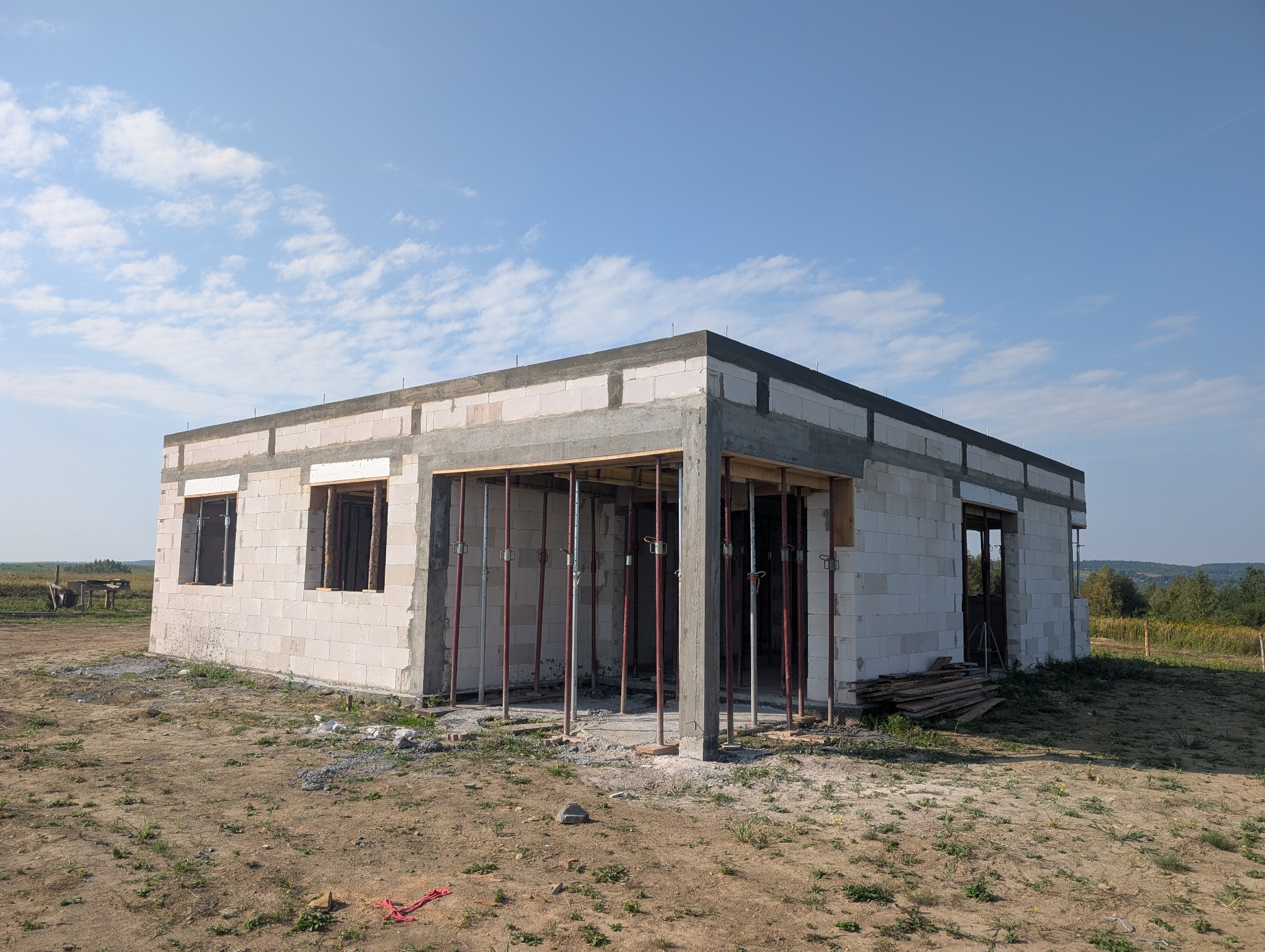 Niedokończony, parterowy dom z białego bloczka, z widocznymi podporami i deskami szalunkowymi, na tle błękitnego nieba z chmurami. Stan surowy otwarty.