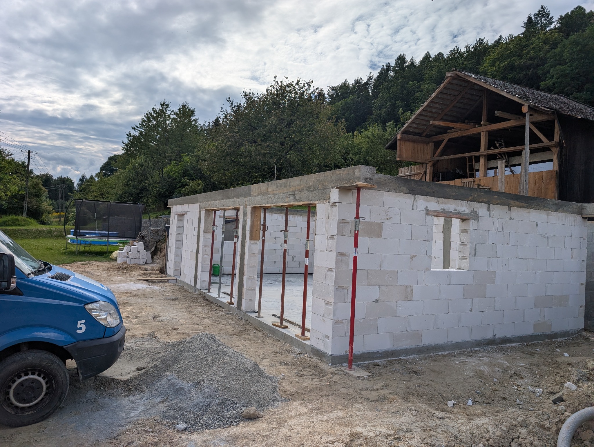 Budowa domu z białych bloczków, widoczne podpory i częściowo wzniesione ściany. W tle las i drewniana konstrukcja. Na pierwszym planie gruz i samochód dostawczy.