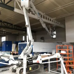 Malowanie konstrukcji stalowej pod sufitem hali produkcyjnej z użyciem podnośnika koszowego, widoczni dwaj pracownicy w kombinezonach malarskich.