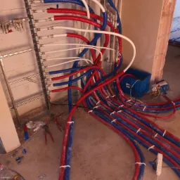 Rozdzielacz instalacji centralnego ogrzewania z rurami PEX w kolorach czerwonym i niebieskim, podłączony do metalowej listwy montażowej, widoczne narzędzia i niebieski pojemnik, surowy stan...