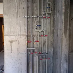 Szare rury instalacji wodnej z czerwonymi i niebieskimi zaworami zamontowane na betonowej ścianie w surowym stanie, widoczne mocowania i fragment niezabudowanego pomieszczenia.