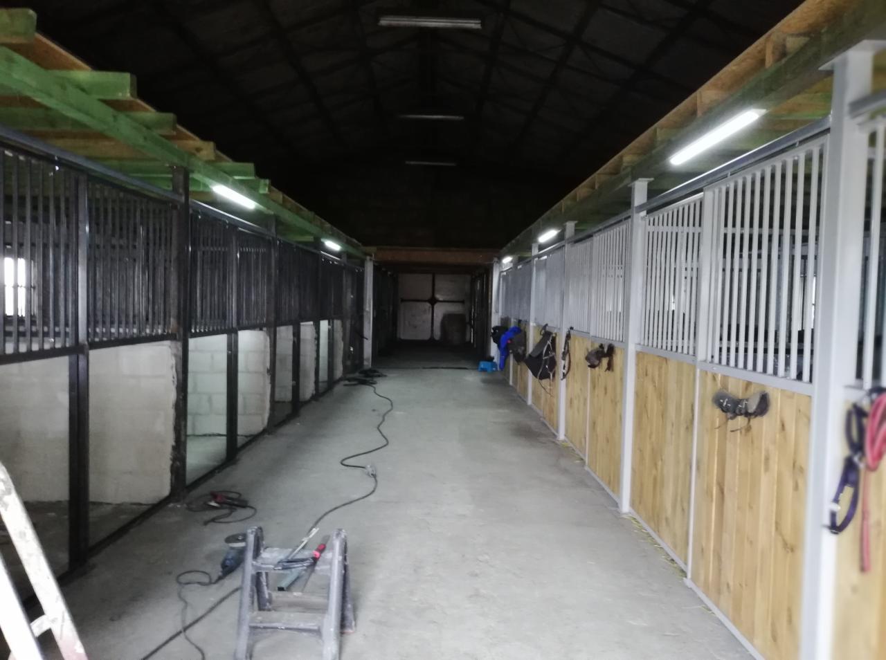 Wnętrze stajni w trakcie budowy lub remontu: widoczne metalowe konstrukcje boksów dla koni, częściowo wykończone drewnem, narzędzia spawalnicze i drabina na posadzce, surowe ściany z bloczków...