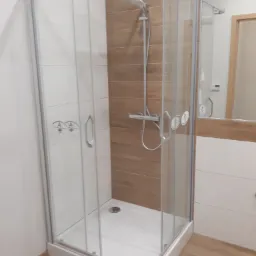 Nowoczesna kabina prysznicowa z przesuwnymi drzwiami, wykończona płytkami imitującymi drewno na ścianie i białymi płytkami na pozostałych ścianach, z widoczną armaturą i odpływem.