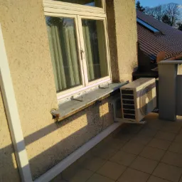 Jednostka zewnętrzna klimatyzatora zamontowana na balkonie, widoczna na tle okna i fragmentu dachu sąsiedniego budynku.