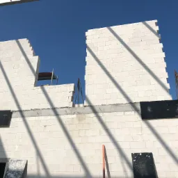 Ściana budynku w trakcie budowy, wzniesiona z białych bloczków, z widocznymi fragmentami konstrukcji stropu i niebieskim niebem w tle.