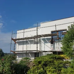 Praca na rusztowaniu: mężczyzna w czarnym podkoszulku i wzorzystych szortach maluje białą elewację domu jednorodzinnego w słoneczny dzień, widok z poziomu ogrodu.