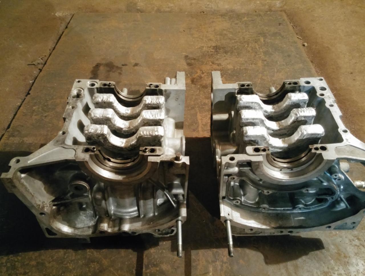 Dwie połówki aluminiowego bloku silnika spalinowego leżące na brudnej metalowej powierzchni, widoczne ślady obróbki i otwory montażowe.