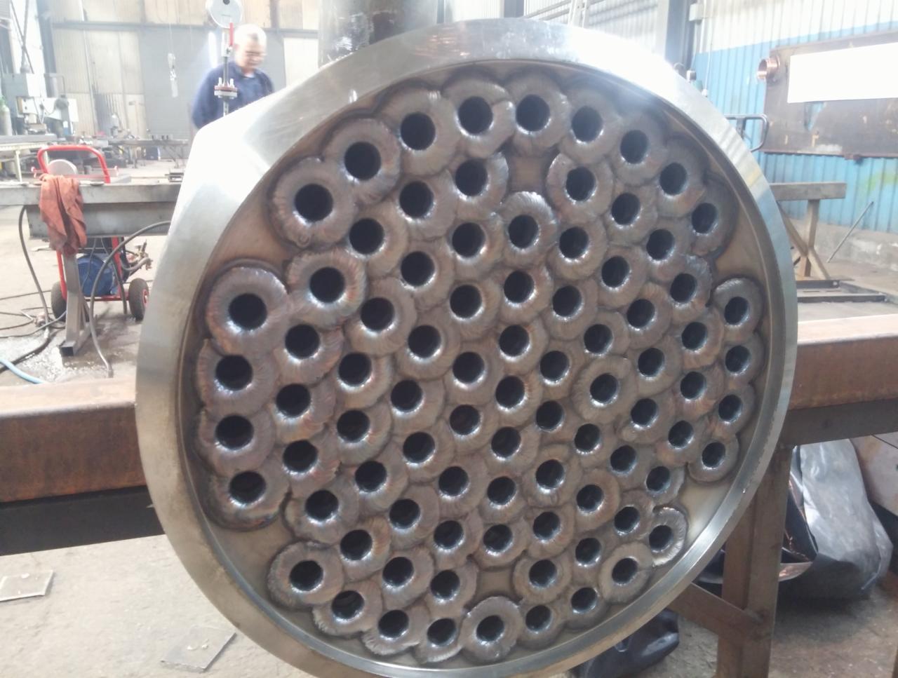 Część wymiennika ciepła z licznymi rurami, po procesie spawania, widoczne ślady spoin, w tle warsztat z pracującym mężczyzną.