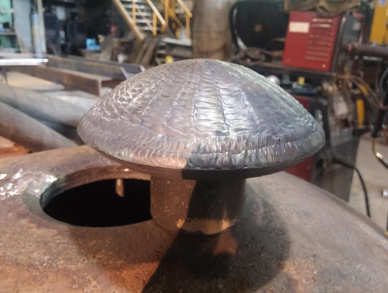 Wyspawana kopuła na metalowym zbiorniku z widocznymi śladami spawania i otworem obok, w tle warsztat z narzędziami.