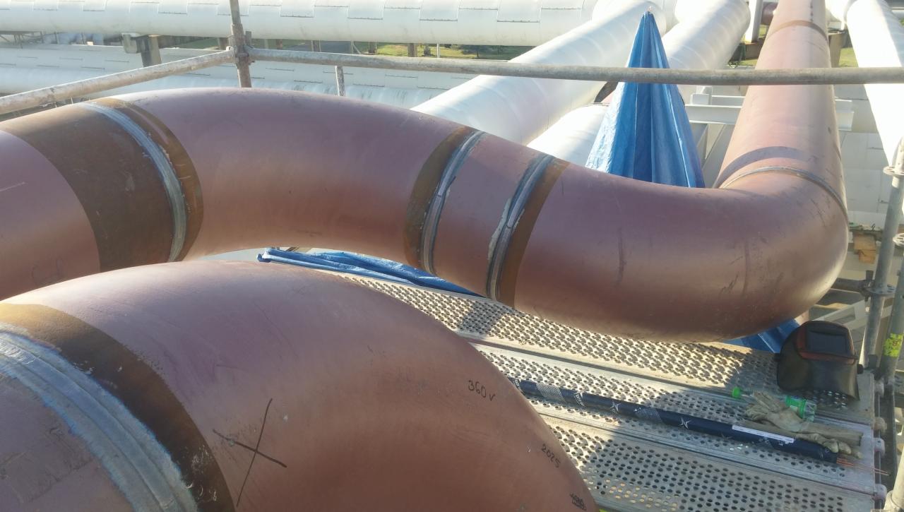 Brązowe rury przemysłowe z widocznymi spawami na rusztowaniu; w tle inne rury i niebieska plandeka, obok leżą rękawice i przyłbica.