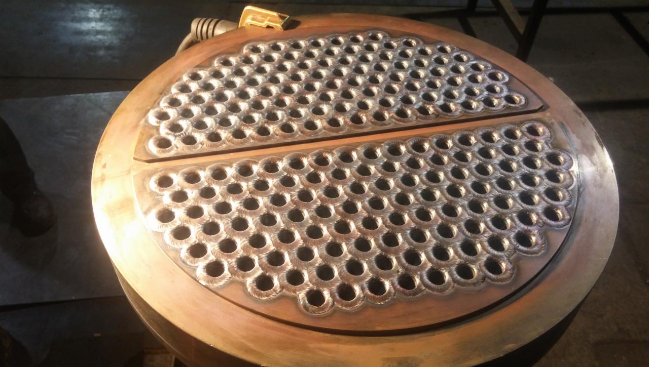 Okrągły element metalowy z licznymi otworami, prawdopodobnie wymiennik ciepła, po obróbce z widocznymi śladami spoin, leżący na metalowej konstrukcji.