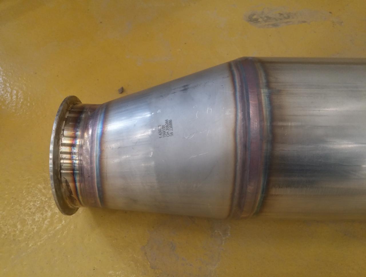 Metalowy element rurowy z widocznymi, precyzyjnymi spawami o charakterystycznym zabarwieniu termicznym, leżący na żółtej powierzchni.