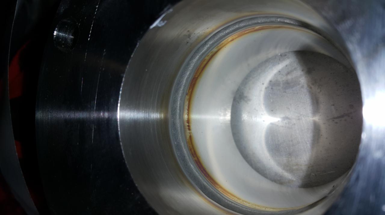 Wnętrze metalowego cylindra z widocznym spoiną obwodową o nieregularnej barwie, od srebrzystej po rdzawą, z ciemniejszymi śladami obróbki na zewnętrznej powierzchni.