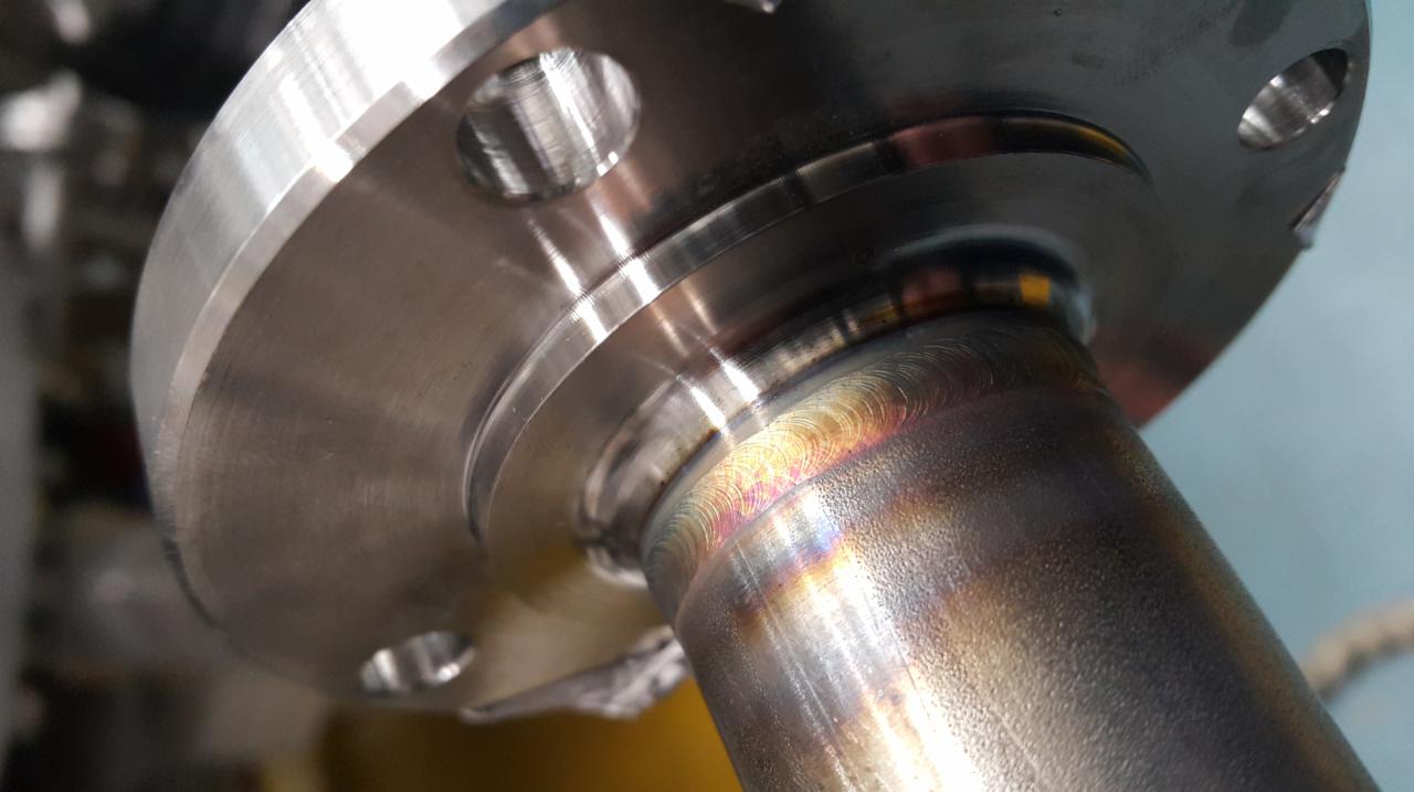 Spoina pachwinowa na metalowej rurze z kołnierzem, widoczne tęczowe przebarwienia po spawaniu.