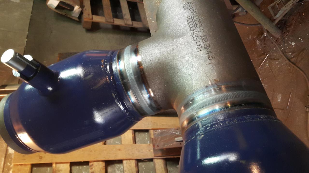 Zespawany trójnik rurowy z widocznymi śladami spawania i niebieską farbą, leżący na drewnianej palecie.