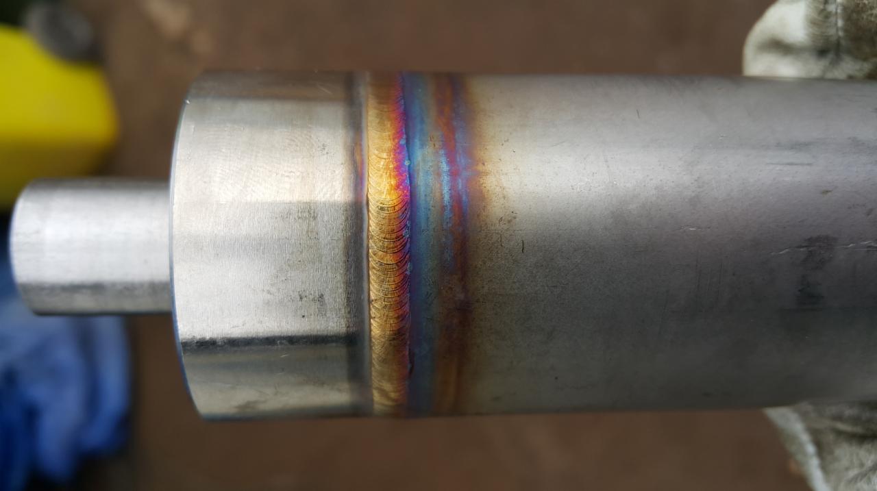 Zespawany metalowy element cylindryczny z widocznymi tęczowymi przebarwieniami na spoinie, trzymany w rękawicy ochronnej.