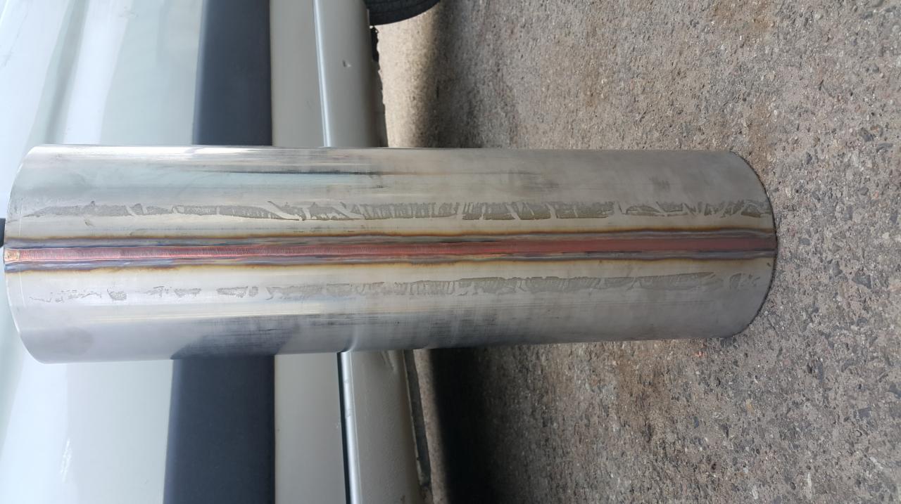 Cylindryczny element ze stali nierdzewnej z widocznym spoiwem spawalniczym na środku, leżący na szorstkiej, betonowej powierzchni, częściowo widoczny bok białego samochodu.