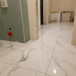 Białe płytki imitujące marmur na podłodze łazienki w trakcie remontu, widoczna zabudowa z płyty gipsowej z pomarańczową rurą, fragment instalacji wodnej i drzwi.