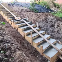 Betonowe schody w drewnianym szalunku w trakcie budowy na zboczu ziemi, widoczna łopata i roślinność w tle.