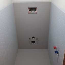 Glazurnik kafelkarz usługi remontowo budowlane - Wnętrze łazienki w trakcie remontu, widoczne płytki z delikatnym wzorem na ścianach, przygotowane przyłącza wodne z zaworami oznaczonymi kolorem czerwonym i niebieskim, otwór na stelaż podtynkowy...