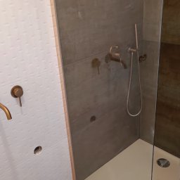 Glazurnik kafelkarz usługi remontowo budowlane - Niedokończona kabina prysznicowa z brązową armaturą, białe płytki z delikatnym wzorem na ścianie obok szarej ściany kabiny, widoczne przewody elektryczne wystające ze ściany.