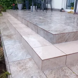 Wykończony taras z ceramicznymi płytkami imitującymi kamień, schody tarasowe z jasnymi płytkami, widok na zadbany ogr&oacute;d i meble ogrodowe.