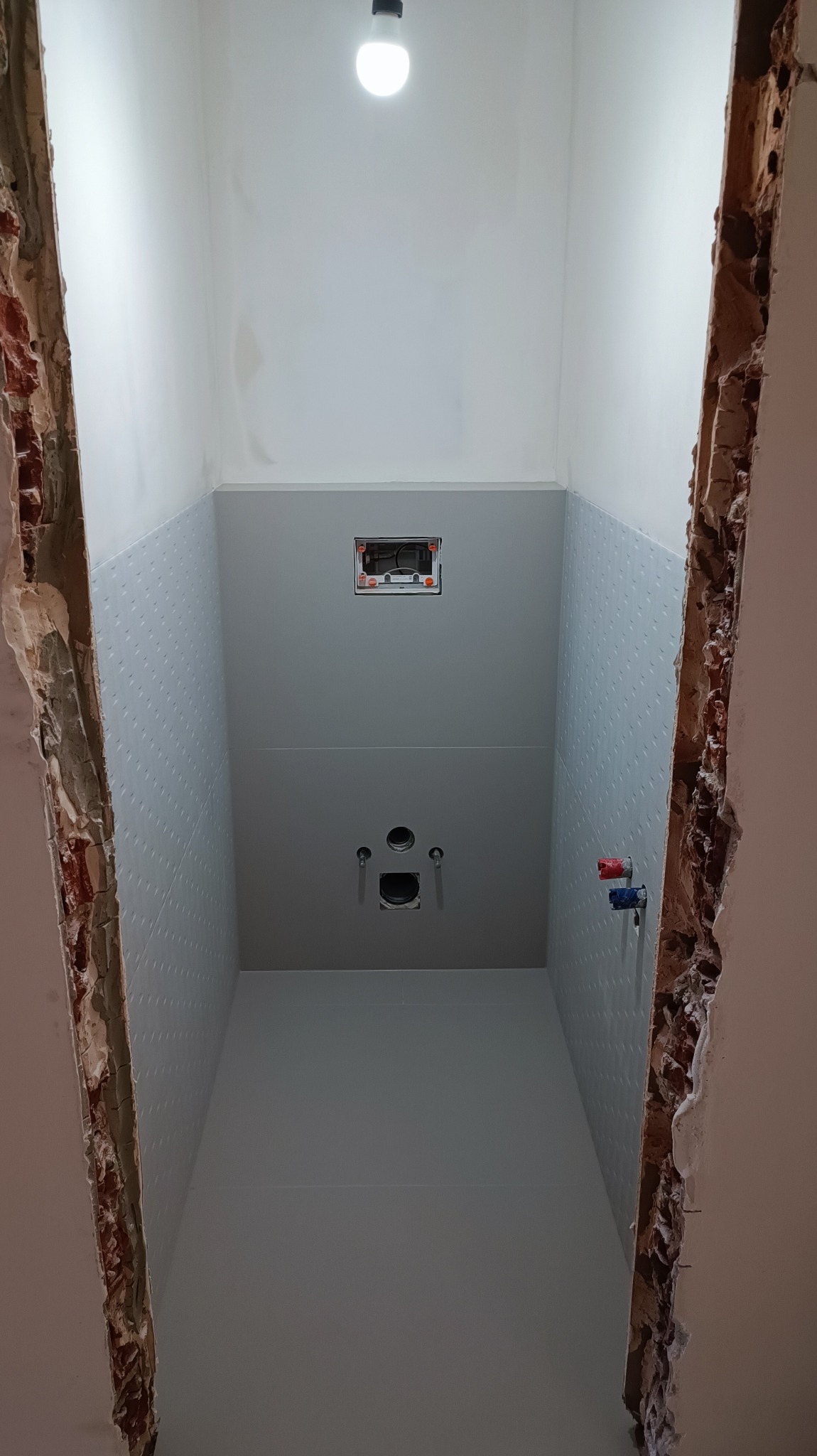 Wnętrze łazienki w trakcie remontu, widoczne przygotowane ściany z częściowo położonymi płytami gipsowymi o delikatnej fakturze, odsłonięta cegła po bokach, instalacja wodna przygotowana do montażu...