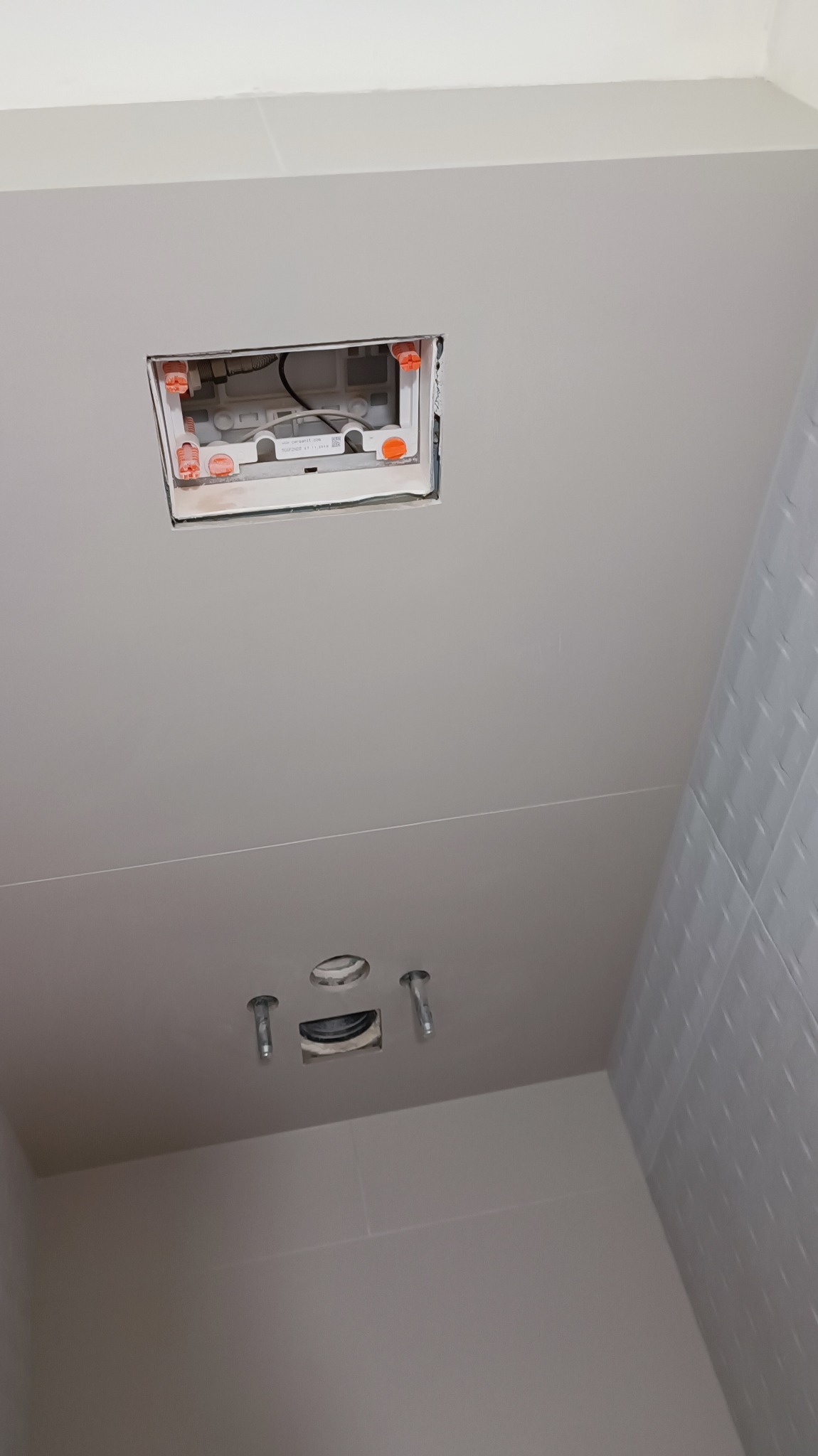 Przygotowanie do montażu stelaża podtynkowego WC: widoczne otwory w ścianie, wystające śruby mocujące i fragment instalacji w ścianie z pomarańczowymi elementami regulacyjnymi, białe płytki...