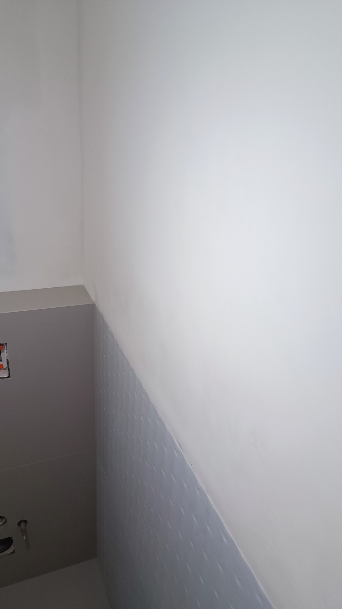 Fragment łazienki z częściowo wyłożonymi szarymi płytkami o subtelnym wzorze, kontrastującymi z białą, gładką ścianą powyżej, w rogu widoczne instalacje hydrauliczne przed zabudową.