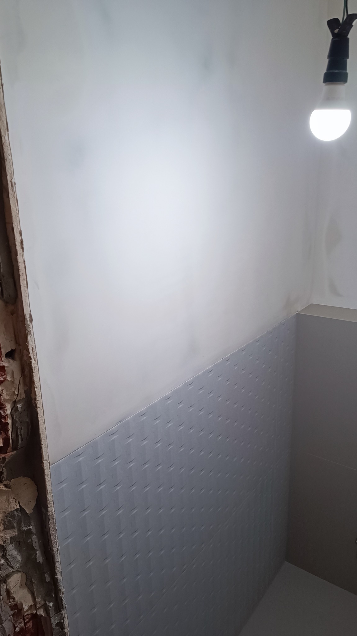 Fragment ściany łazienki z częściowo położonymi szarymi płytkami o wzorze geometrycznym, widoczny surowy fragment ściany z cegły i wisząca żarówka.