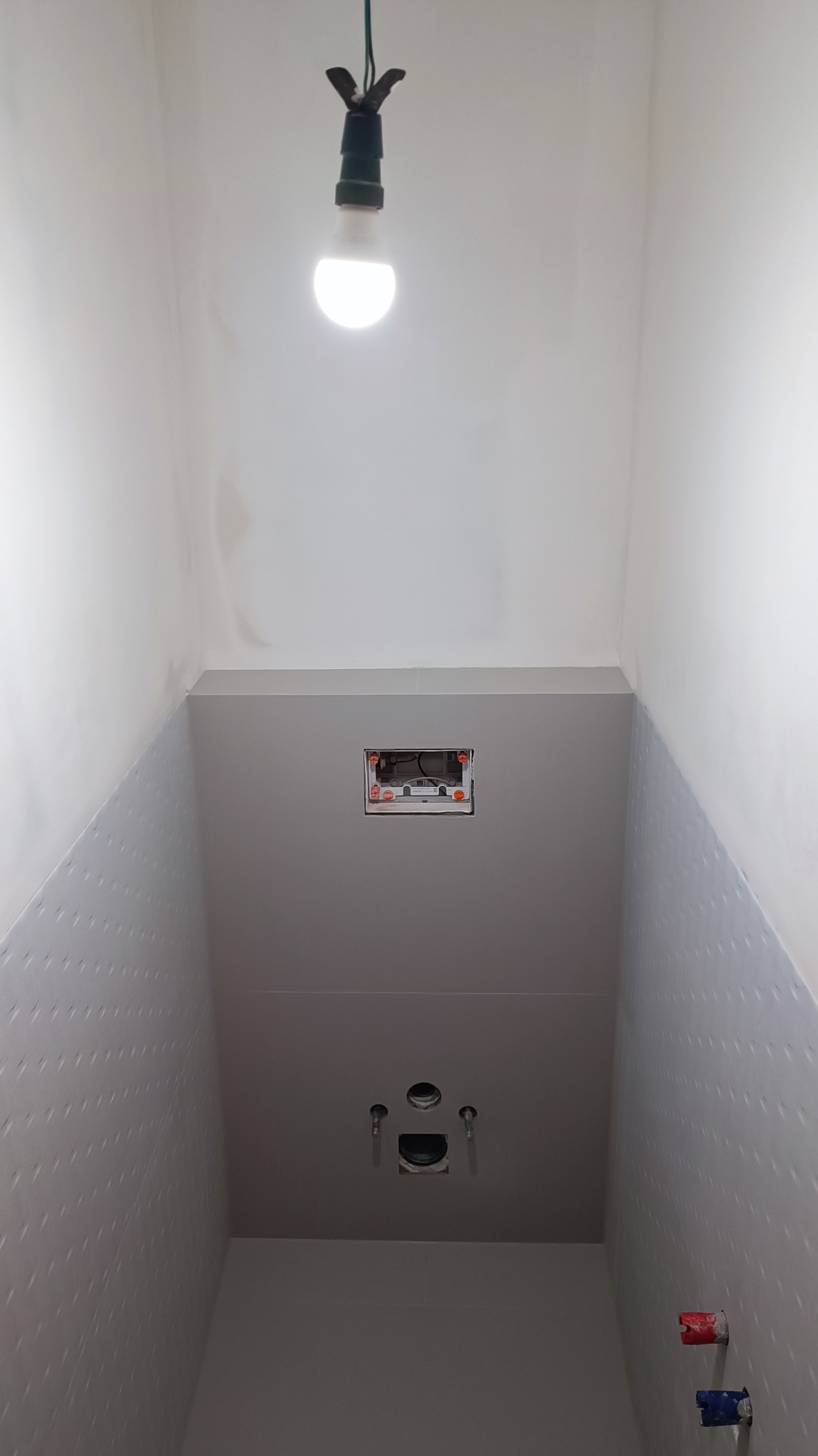 Wnętrze łazienki w trakcie remontu, widoczne płytki z delikatnym wzorem na ścianach, przygotowane instalacje wodno-kanalizacyjne, otwór na puszkę elektryczną i prowizoryczne oświetlenie w postaci...