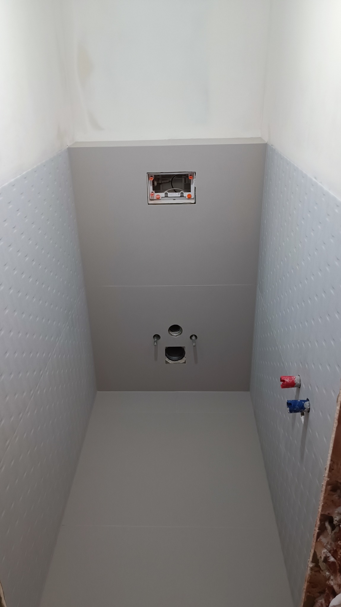 Wnętrze łazienki w trakcie remontu, widoczne płytki z delikatnym wzorem na ścianach, przygotowane przyłącza wodne z zaworami oznaczonymi kolorem czerwonym i niebieskim, otwór na stelaż podtynkowy...