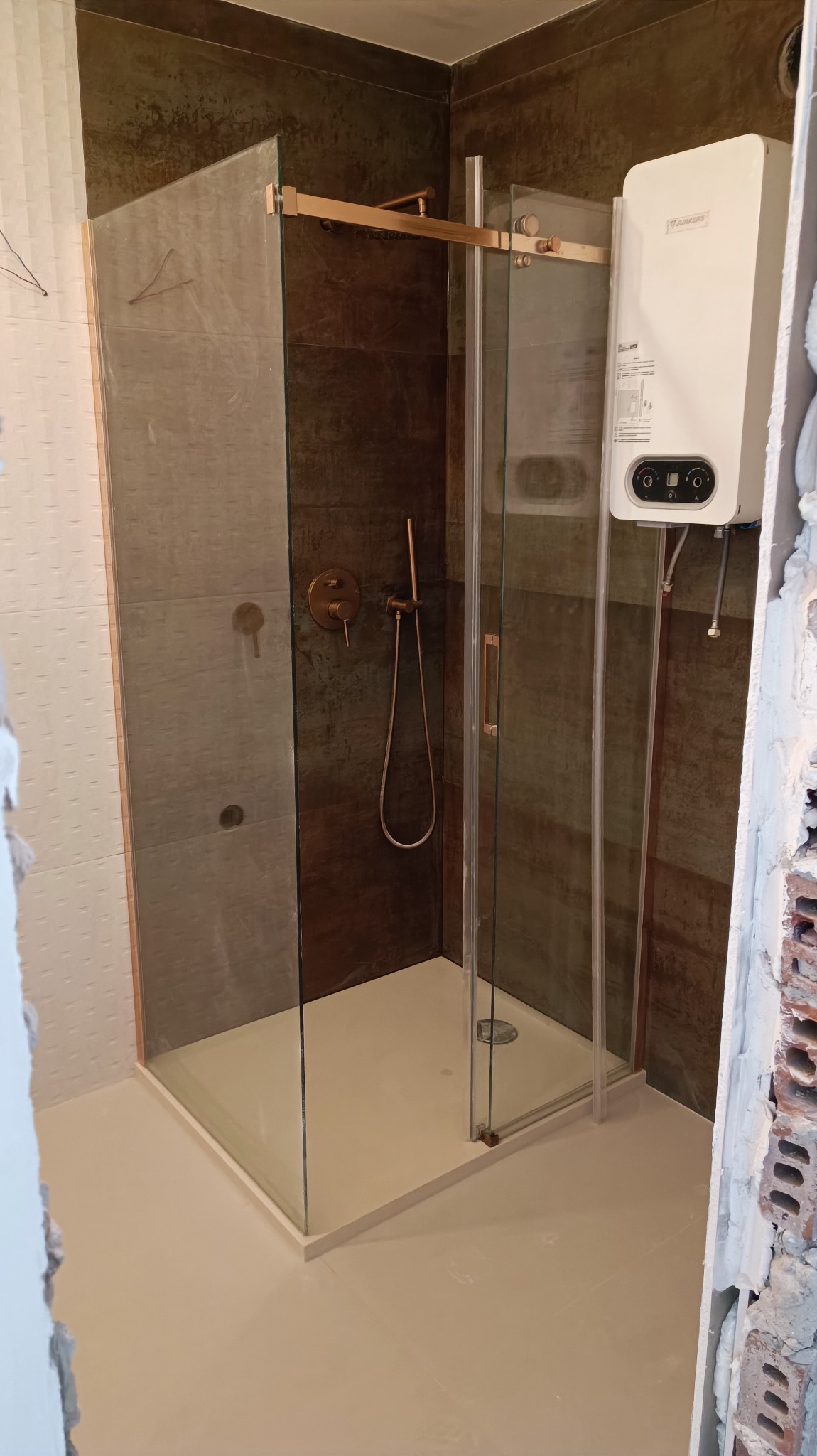 Nowoczesna kabina prysznicowa z przesuwnymi drzwiami, armaturą w kolorze miedzi i płytkami imitującymi rdzę, z zamontowanym bojlerem gazowym na ścianie, widok na wykończoną podłogę i fragment...