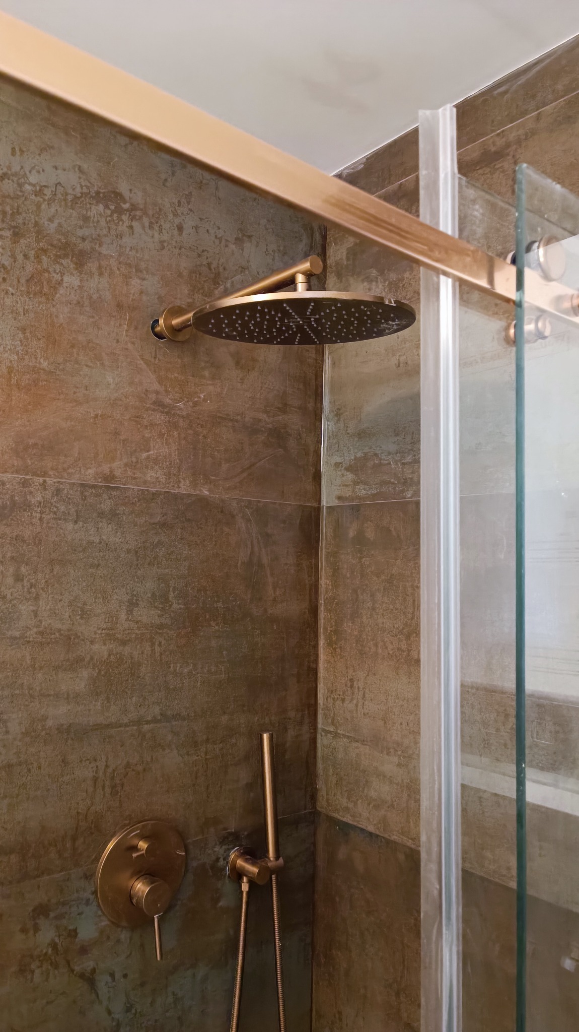 Nowoczesna kabina prysznicowa z deszczownicą w kolorze miedzianym, zamontowana na ścianie wyłożonej płytkami imitującymi beton, z widoczną szklaną ścianą kabiny.